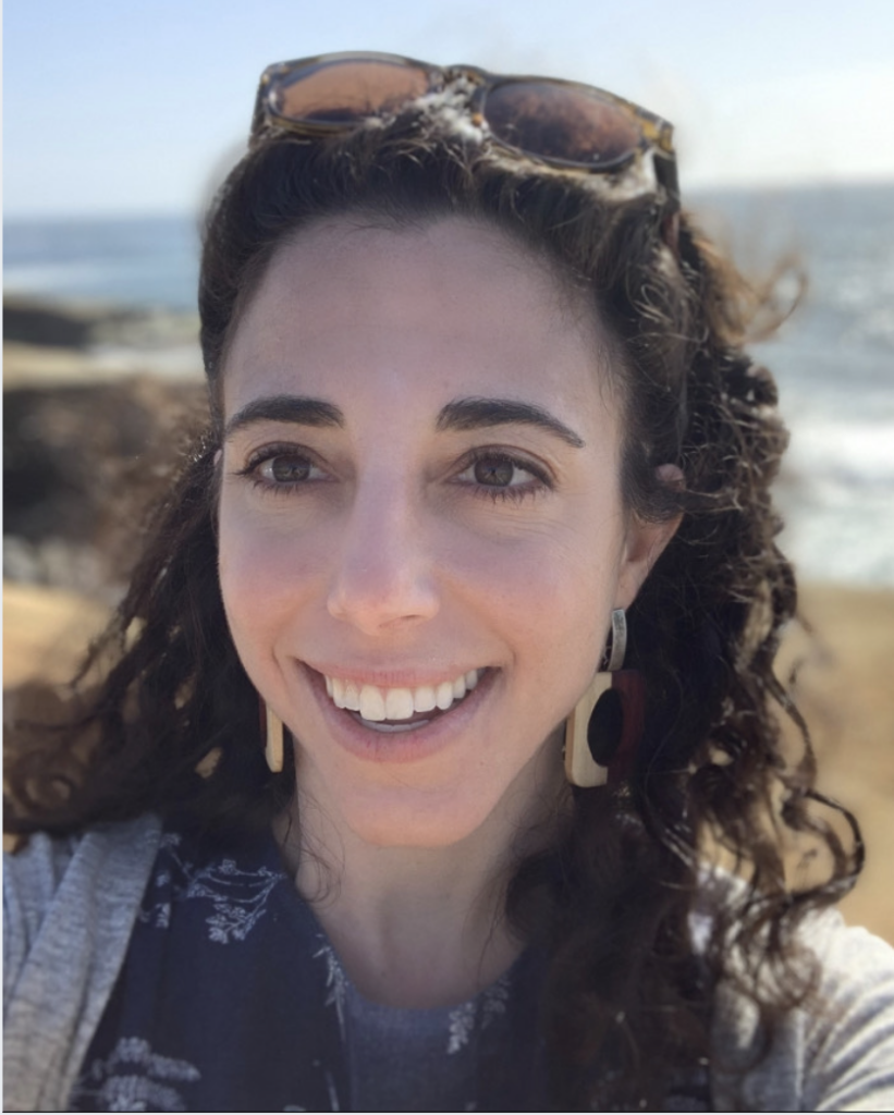 A woman with curly hair and sunglasses on her head smiles outdoors near a body of water.